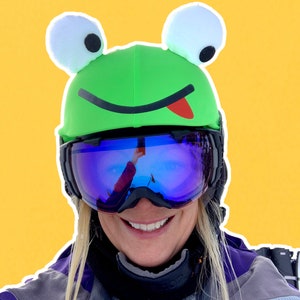 Pode incluir: Um capacete de esqui verde em forma de sapo com uma l&iacute;ngua vermelha e grandes olhos brancos. O capacete tem uma viseira preta com lentes azuis.