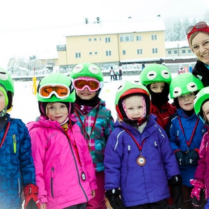 Pode incluir: Um grupo de crian&ccedil;as usando capacetes de sapo verdes e roupas de esqui est&atilde;o em uma &aacute;rea com neve. Todos est&atilde;o sorrindo e segurando medalhas. Uma mulher adulta usando um chap&eacute;u vermelho e roupas de esqui est&aacute; atr&aacute;s deles.