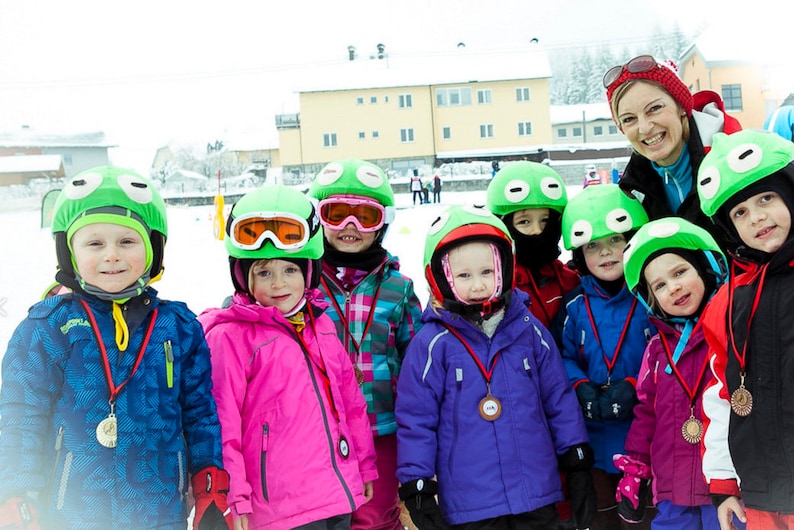 Pode incluir: Um grupo de crian&ccedil;as usando capacetes de sapo verdes e roupas de esqui est&atilde;o em uma &aacute;rea com neve. Todos est&atilde;o sorrindo e segurando medalhas. Uma mulher adulta usando um chap&eacute;u vermelho e roupas de esqui est&aacute; atr&aacute;s deles.