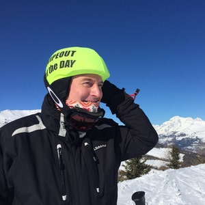 Puede incluir: Una persona lleva un casco de esquí amarillo brillante con el texto "Wipeout of the Day" en él. La persona lleva una chaqueta de esquí negra con una franja blanca en el hombro y un cuello de lana rojo y blanco con estampado. La persona está de pie en un paisaje montañoso nevado.