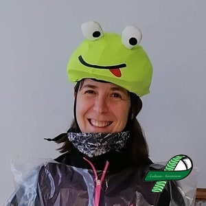 Pode incluir: Uma mulher usando um capacete verde em forma de sapo com uma l&iacute;ngua vermelha e um len&ccedil;o com padr&atilde;o preto e branco. Ela est&aacute; usando um poncho de pl&aacute;stico transparente e uma fita verde com o texto "Scoliosis Awareness".
