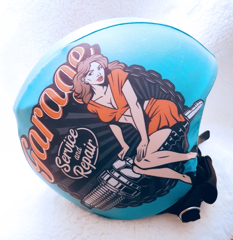 May include: A blue helmet cover with a vintage-style illustration of a woman sitting on a spark plug. The text "Garage, Service and Repair" is printed on the cover.