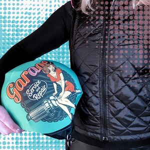 May include: A teal blue helmet cover with a vintage pinup girl design. The cover features the text "Garage Service and Repair" in a retro font.