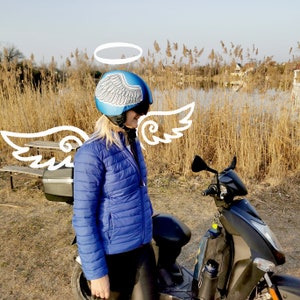 May include: A woman wearing a blue helmet with white angel wings painted on it, standing next to a black scooter. The helmet has a halo drawn above it.