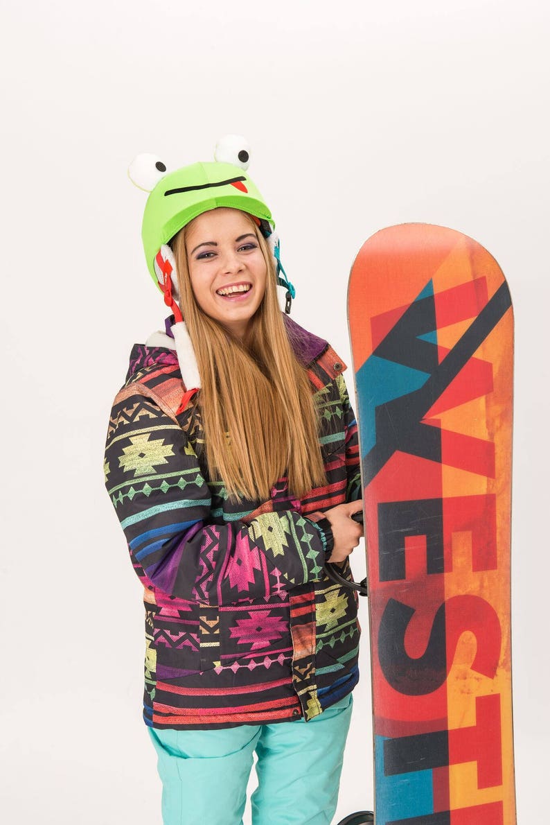 Pode incluir: Uma mulher vestindo uma jaqueta estampada colorida e cal&ccedil;as turquesa est&aacute; segurando um snowboard. Ela est&aacute; usando um capacete de sapo verde com uma l&iacute;ngua vermelha.