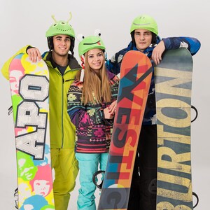 Pode incluir: Tr&ecirc;s amigos usando capacetes coloridos e segurando pranchas de snowboard. As pranchas de snowboard t&ecirc;m designs e logotipos diferentes, incluindo "APO", "YES" e "BURTON".