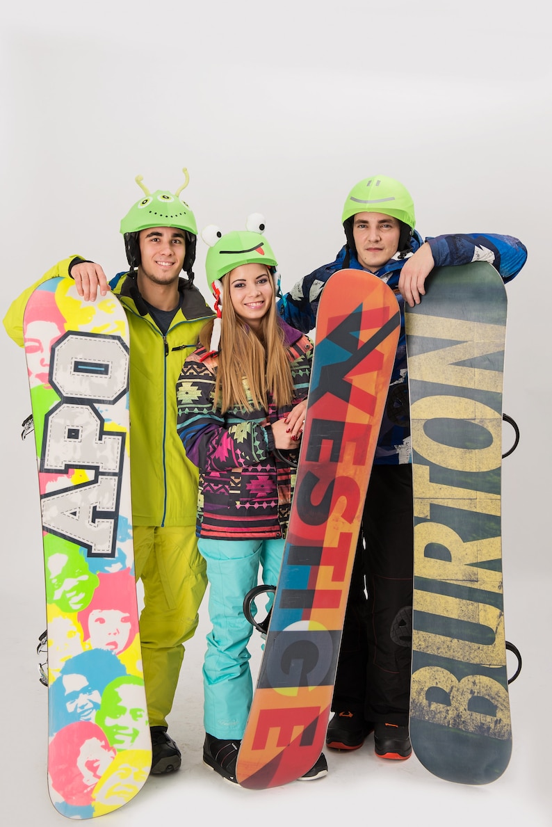 Pode incluir: Tr&ecirc;s amigos usando capacetes coloridos e segurando pranchas de snowboard. As pranchas de snowboard t&ecirc;m designs e logotipos diferentes, incluindo "APO", "YES" e "BURTON".