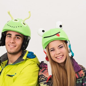 Pode incluir: Duas pessoas usando capacetes verdes com divertidos desenhos de animais. O capacete &agrave; esquerda tem um design de alien&iacute;gena verde com antenas. O capacete &agrave; direita tem um design de sapo verde com a l&iacute;ngua de fora. Ambas as pessoas est&atilde;o sorrindo.