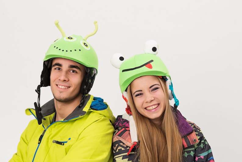 Pode incluir: Duas pessoas usando capacetes verdes com divertidos desenhos de animais. O capacete &agrave; esquerda tem um design de alien&iacute;gena verde com antenas. O capacete &agrave; direita tem um design de sapo verde com a l&iacute;ngua de fora. Ambas as pessoas est&atilde;o sorrindo.