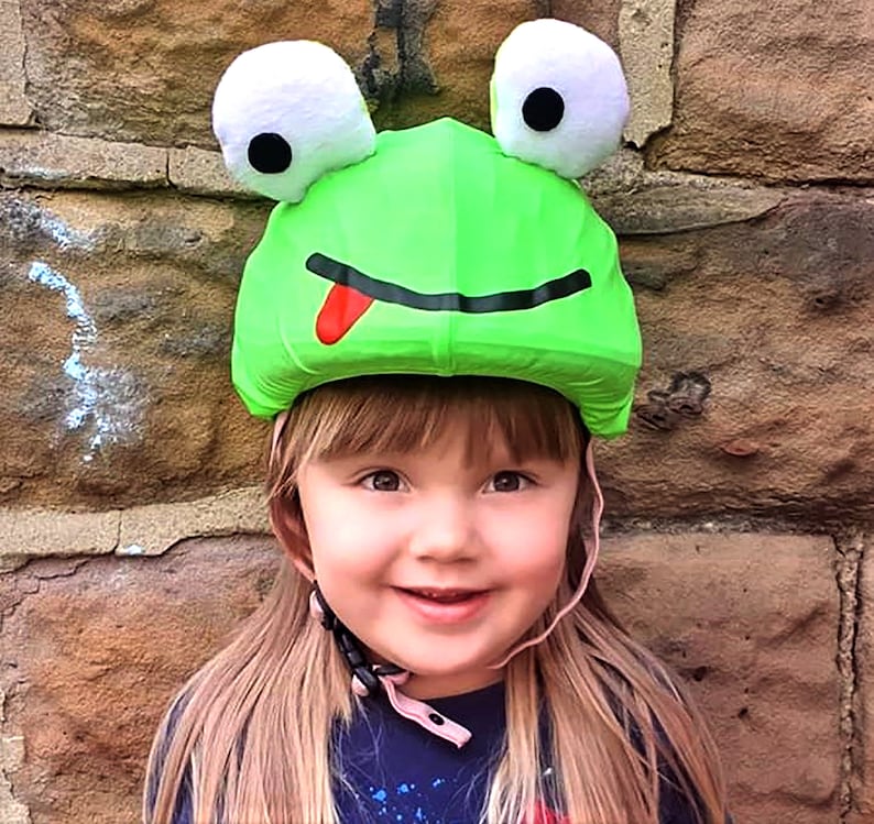 Pode incluir: Um capacete de r&atilde; verde com uma l&iacute;ngua vermelha e olhos brancos. O capacete est&aacute; sendo usado por uma crian&ccedil;a com cabelo longo e loiro.