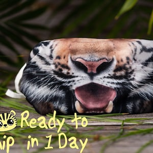 May include: A black, white, and orange tiger face mask with a white elastic band. The mask is made of fabric and has a realistic tiger print. The tiger's mouth is open and its tongue is sticking out. The mask is lying on a wooden surface with green grass in the background.