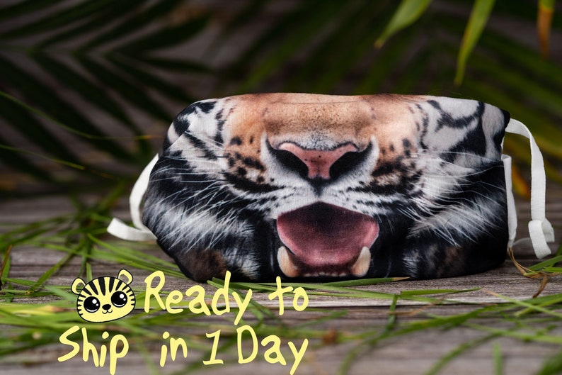 May include: A black, white, and orange tiger face mask with a white elastic band. The mask is made of fabric and has a realistic tiger print. The tiger's mouth is open and its tongue is sticking out. The mask is lying on a wooden surface with green grass in the background.