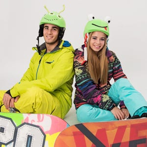 Pode incluir: Duas pessoas usando capacetes coloridos com designs divertidos est&atilde;o sentadas em snowboards. A pessoa &agrave; esquerda est&aacute; usando um macac&atilde;o de neve amarelo e a pessoa &agrave; direita est&aacute; usando um macac&atilde;o de neve estampado em azul e preto.
