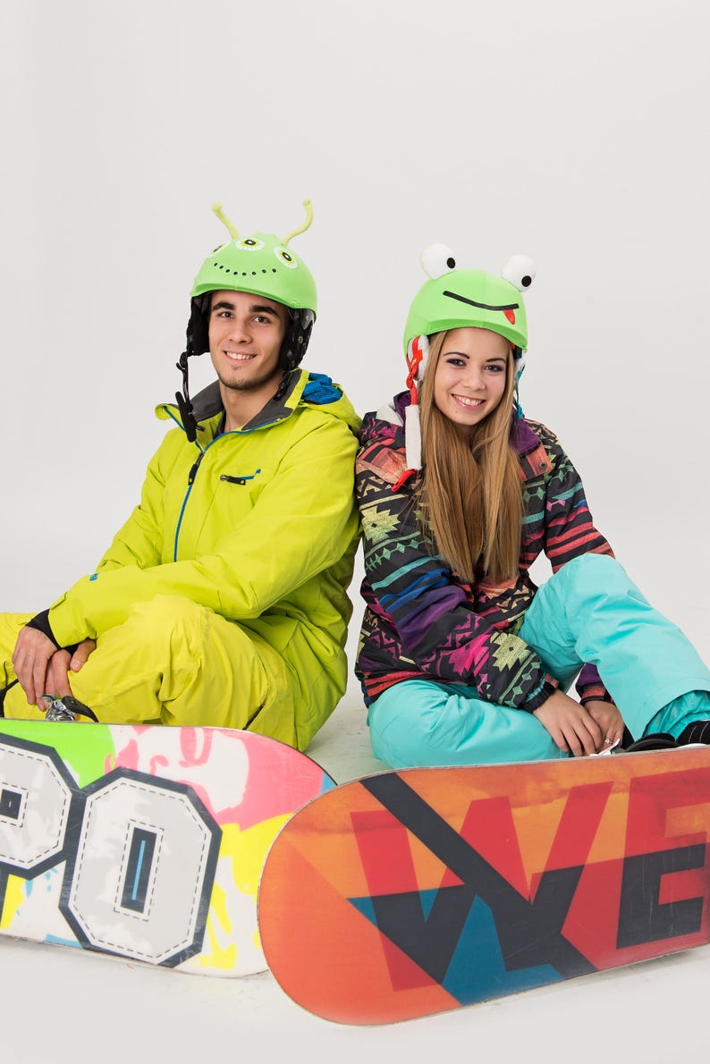 Pode incluir: Duas pessoas usando capacetes coloridos com designs divertidos est&atilde;o sentadas em snowboards. A pessoa &agrave; esquerda est&aacute; usando um macac&atilde;o de neve amarelo e a pessoa &agrave; direita est&aacute; usando um macac&atilde;o de neve estampado em azul e preto.