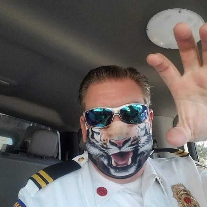 May include: A man wearing a white uniform with a tiger face mask and blue sunglasses. He is in a car and has his hand raised.