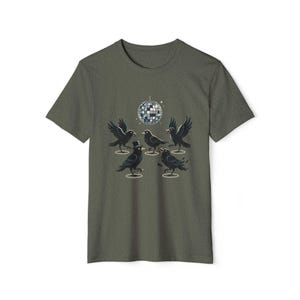 May include: An olive green short-sleeve t-shirt featuring a graphic print of five black crows dancing under a silver disco ball. One crow wears a small black top hat, and each bird is positioned within a thin white circle on the fabric.