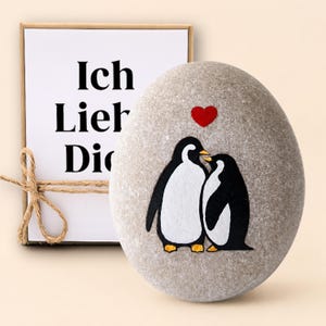 Könnte beinhalten: Ein glatter, ovaler grauer Stein mit einer handgemalten Illustration von zwei Pinguinen, die sich gegenüberstehen, mit einem kleinen roten Herz darüber, begleitet von einer rechteckigen weißen Karte mit der Aufschrift Ich Lieb Dich und Bindfaden.