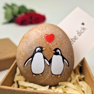 Könnte beinhalten: Bemalter Stein mit zwei Pinguinen, die sich an den Händen halten, darüber ein rotes Herz. Die Pinguine sind schwarz und weiß mit gelben Schnäbeln und Füßen. Der Stein befindet sich in einer braunen Schachtel mit Papier.