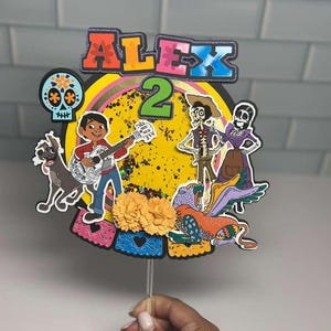 May include: A colorful cake topper featuring characters from the movie Coco. The topper includes the name "ALEX" and the number "2". The design incorporates a yellow background, a guitar-playing boy, and other animated figures. The topper is held by a clear stick.