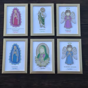 May include: Six framed religious art pieces. Each features a cutout design of a saint or angel in various colors, including pink, green, and blue, set against a white background. The frames are gold-colored, and each piece has text in Spanish.