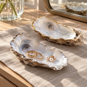 May include: Two decorative oyster shell dishes with iridescent white interiors and gold-painted edges. One shell holds a gold band ring, a thin diamond-encrusted ring, and a pair of small white pearl stud earrings.