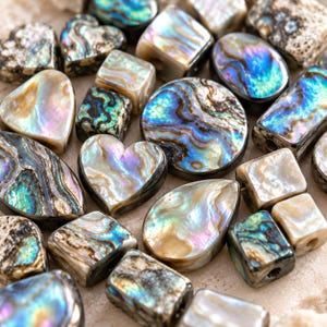 Op de afbeelding: Een verzameling diverse abalone schelpkralen in verschillende vormen, waaronder harten, vierkanten, cirkels en druppels. Elke kraal heeft een gepolijst, iriserend oppervlak met wervelende patronen in blauwe, groene, paarse en crèmekleurige tinten.
