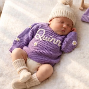 May include: A sleeping baby wearing a lavender sweater with the name "Quinn" in white script. The sweater is adorned with small daisy appliqués. The baby also wears a cream-colored knit hat with a pom-pom and matching socks. A lavender knit hat is next to the baby.