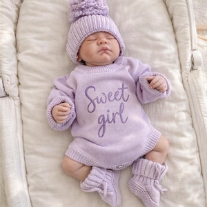 May include: A baby wearing a lavender knit outfit, including a hat with a pom-pom, a long-sleeved romper with "Sweet girl" embroidered on the front, and matching booties. The baby is lying on a white surface.