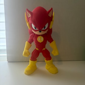 May include: A red and yellow action figure of a hybrid character, combining the features of Sonic the Hedgehog and The Flash. The figure has yellow boots, gloves, and lightning bolt accents, with a Flash emblem on the chest.