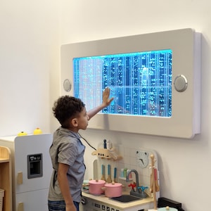 May include: A white rectangular sensory bubble wall with blue illuminated bubbles. A child is interacting with the wall, touching the surface. The wall is mounted on a white wall. A toy kitchen set is in the foreground.