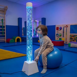 May include: A tall, white sensory bubble tube with blue lights and bubbles. A child sits on a blue exercise ball, interacting with the light-up tube. The room has blue flooring and walls, with colorful play equipment.