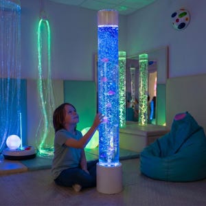 May include: A sensory room with a tall, illuminated bubble tube filled with blue water and colorful, floating objects. A child sits nearby, interacting with the tube. Other sensory elements include fiber optic lights and a beanbag chair.