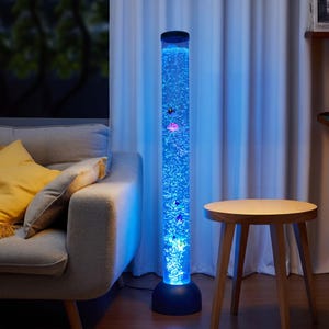 May include: A tall, cylindrical bubble lamp with a blue illuminated interior. The lamp features simulated jellyfish and fish, creating a calming ambiance. The lamp is placed next to a gray sofa and a small wooden side table.