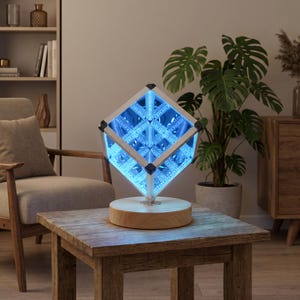 May include: A cube-shaped infinity mirror lamp featuring a bright blue LED light display. The structure is supported by a light-colored wooden circular base, creating a geometric visual effect with repeating light patterns inside the frame.