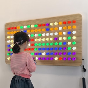 May include: A wooden board with a grid of colorful, illuminated buttons. The buttons are arranged in rows and columns, displaying a spectrum of colors. A child is standing in front of the board, interacting with it. The board is mounted on a white wall.