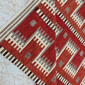 May include: A red and white geometric patterned rug with a diamond design. The rug features a border with stripes and a repeating pattern of white, gray, and red shapes. The rug is made of woven fabric.