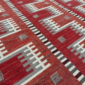 May include: A red and white patterned rug with geometric designs. The rug features a repeating pattern of white and gray shapes against a red background. The rug's texture is visible, suggesting a woven construction.