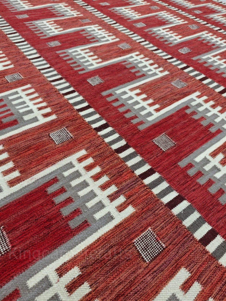 May include: A red and white patterned rug with geometric designs. The rug features a repeating pattern of white and gray shapes against a red background. The rug's texture is visible, suggesting a woven construction.