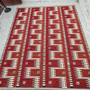 May include: A rectangular rug with a geometric pattern in shades of red, white, and gray. The design features repeating rectangular shapes and lines. The rug has a cream-colored border and is placed on a light-colored tiled floor.