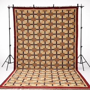 May include: A large, rectangular woven rug with a geometric pattern. The rug features a tan background with a repeating pattern of brown squares and black cross shapes, accented with small red dots. The border is a solid dark red. The rug is displayed on a white background.