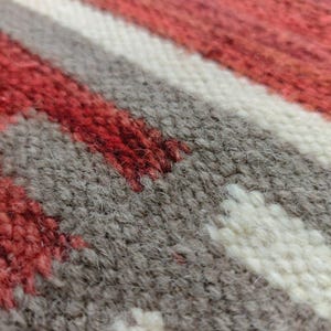 May include: Close-up of a woven textile with a geometric pattern. The design features red, white, and gray stripes and shapes. The texture of the fabric is visible, with individual threads creating a textured surface.