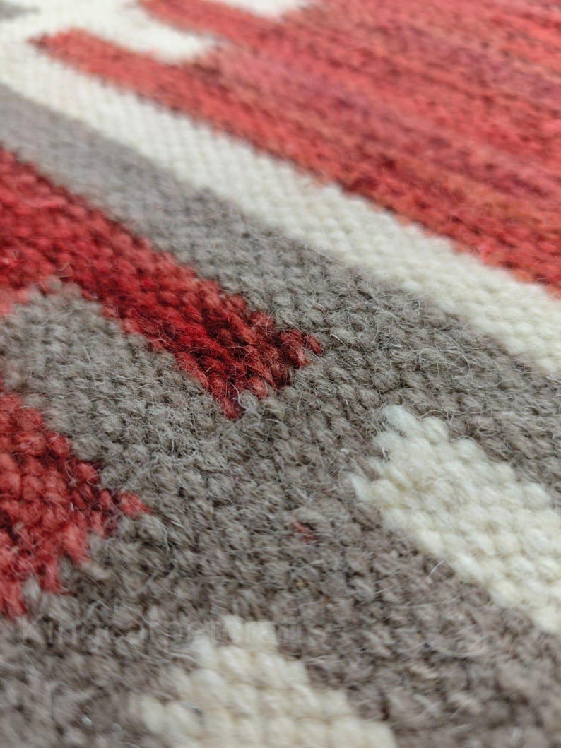 May include: Close-up of a woven textile with a geometric pattern. The design features red, white, and gray stripes and shapes. The texture of the fabric is visible, with individual threads creating a textured surface.