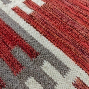 May include: Close-up of a woven textile with a geometric pattern. The design features alternating bands of red, white, and gray. The texture of the fabric is visible, with a slightly rough, handmade appearance. The color palette is warm and earthy.