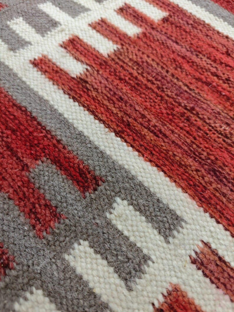 May include: Close-up of a woven textile with a geometric pattern. The design features alternating bands of red, white, and gray. The texture of the fabric is visible, with a slightly rough, handmade appearance. The color palette is warm and earthy.