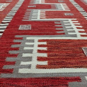 May include: A red, white, and gray geometric patterned rug. The rug features a repeating design of rectangles and lines in varying shades of red, white, and gray. The rug's texture appears to be woven.