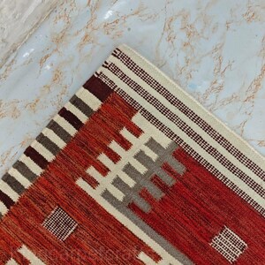 May include: A corner of a woven rug with a geometric pattern. The rug features a color palette of red, cream, gray, and brown. The design includes rectangular and striped elements, creating a textured appearance. The rug is placed on a light-colored surface.