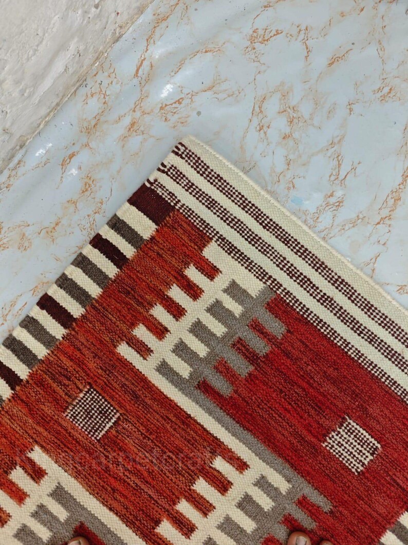 May include: A corner of a woven rug with a geometric pattern. The rug features a color palette of red, cream, gray, and brown. The design includes rectangular and striped elements, creating a textured appearance. The rug is placed on a light-colored surface.