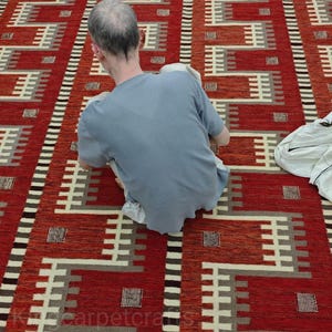May include: A red and cream-colored geometric patterned rug. The rug features a repeating pattern of rectangular shapes and lines in shades of red, cream, and gray. The rug is rectangular and appears to be made of woven material.