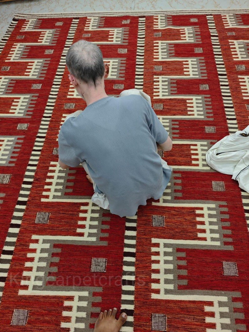 May include: A red and cream-colored geometric patterned rug. The rug features a repeating pattern of rectangular shapes and lines in shades of red, cream, and gray. The rug is rectangular and appears to be made of woven material.