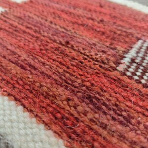 May include: Close-up of a woven rug with a geometric pattern. The rug features a blend of red, orange, and brown hues, with white and gray accents. The texture of the rug is visible, showcasing the woven fibers.